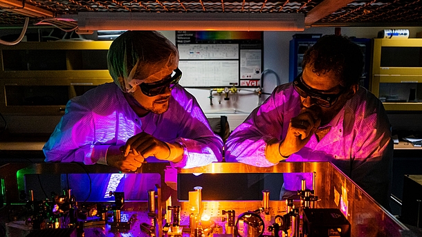 2 people in protective gowns and goggles are standing in a light laboratory looking at an optical experiment with lasers that shine in orange-violet colours.