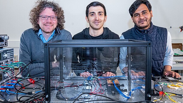 Three researchers in casual clothes stand behind an optical table in the laser laboratory of Leibniz Universität Hannover, in front of them is a photonic chip in a glass box.
