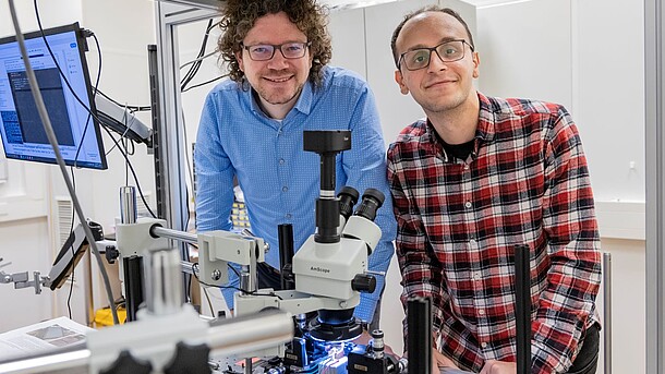 Two scientists stand in a laser laboratory behind an optical table with a large grey-black microscope.