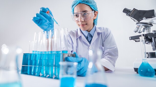 Woman in lab coat and blue protective hat and blue rubber gloves sits in front of a row of test tubes with blue liquid.