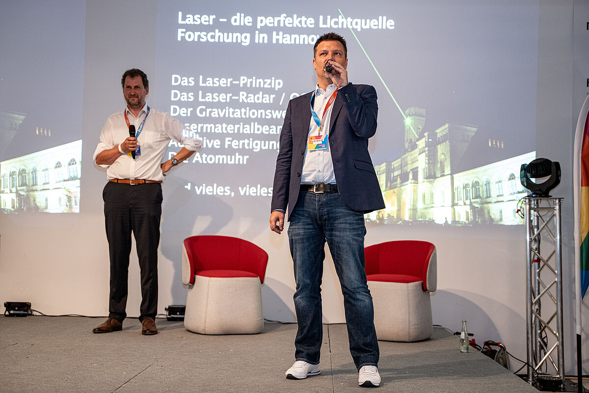Zwei Männer halten Mikrofone in den Händen und stehen auf einer Bühne.